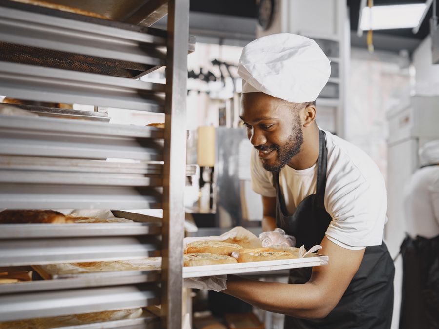 A smiling baker holding a tray of fresh baked goods in a bakery kitchen